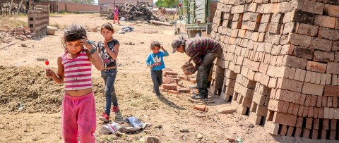 Tres niñas comen una paleta mientras su padre, fabricante de ladrillos, trabaja cerca.